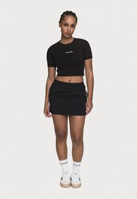 Svart avkortet t-skjorte med logo, svart skjørt med snøring, hvite sokker med logo, og svart-hvite sneakers med beige såle.