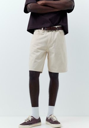 Személy bézs rövidnadrágot, barna övet, sötétbarna inget, fehér zoknit és barna sportcipőt visel, karját keresztbe téve áll.