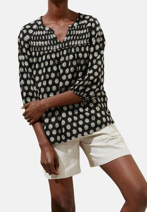Femme portant un chemisier noir à manches longues avec un motif floral blanc et un short beige, debout les bras croisés devant un fond blanc.