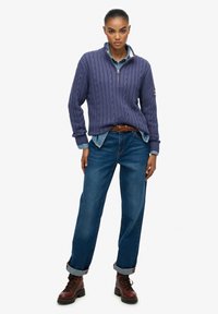 Suéter de punto cableado morado con un cuello de media cremallera, superpuesto a una camisa de mezclilla. Combinado con jeans azules con puños enrollados y botas marrones con cordones.