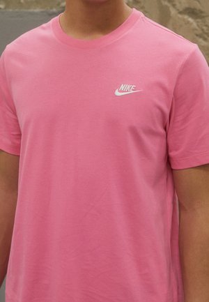 Personne portant un t-shirt Nike rose vif avec un petit logo blanc sur la poitrine gauche, debout devant un mur en pierre neutre.