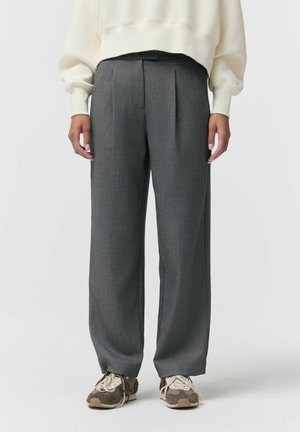 Osoba ubrana w szerokie, szare spodnie, beżowo-brązowe sneakersy i kremową bluzkę z długim rękawem, stojąca na tle jednolitej białej ściany.