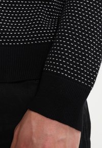 Maglione nero lavorato a maglia con puntini bianchi, con polsini a coste. La trama è liscia, con un design aderente e dettagli sottili.