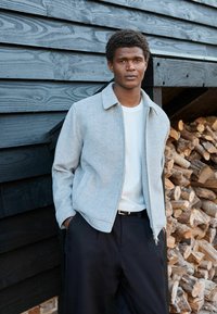 Grijze gezoomde jas van zacht materiaal, witte T-shirt met ronde hals, zwarte broeken met een zachte textuur en een relaxed fit. Houten stammen op de achtergrond.