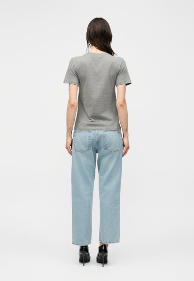 Γυναίκα με μακριά καστανά μαλλιά που φοράει γκρι μπλουζάκι, ανοιχτό μπλε τζιν με ίσια πόδια και μαύρα παπούτσια με ψηλό τακούνι, ακουμπώντας από πίσω.