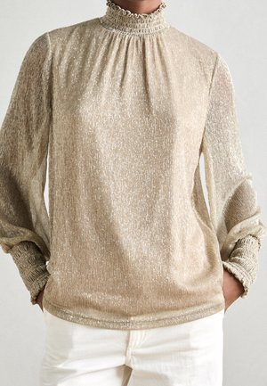 Person iført en skinnende beige bluse med lange ærmer, med bølgende høj hals og manchetter, kombineret med hvide bukser, hænderne i lommerne.