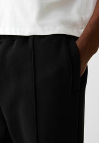 Black sweatpants feature a smooth texture, elastic waistband, and side pockets, paired with a white t-shirt partially visible above.