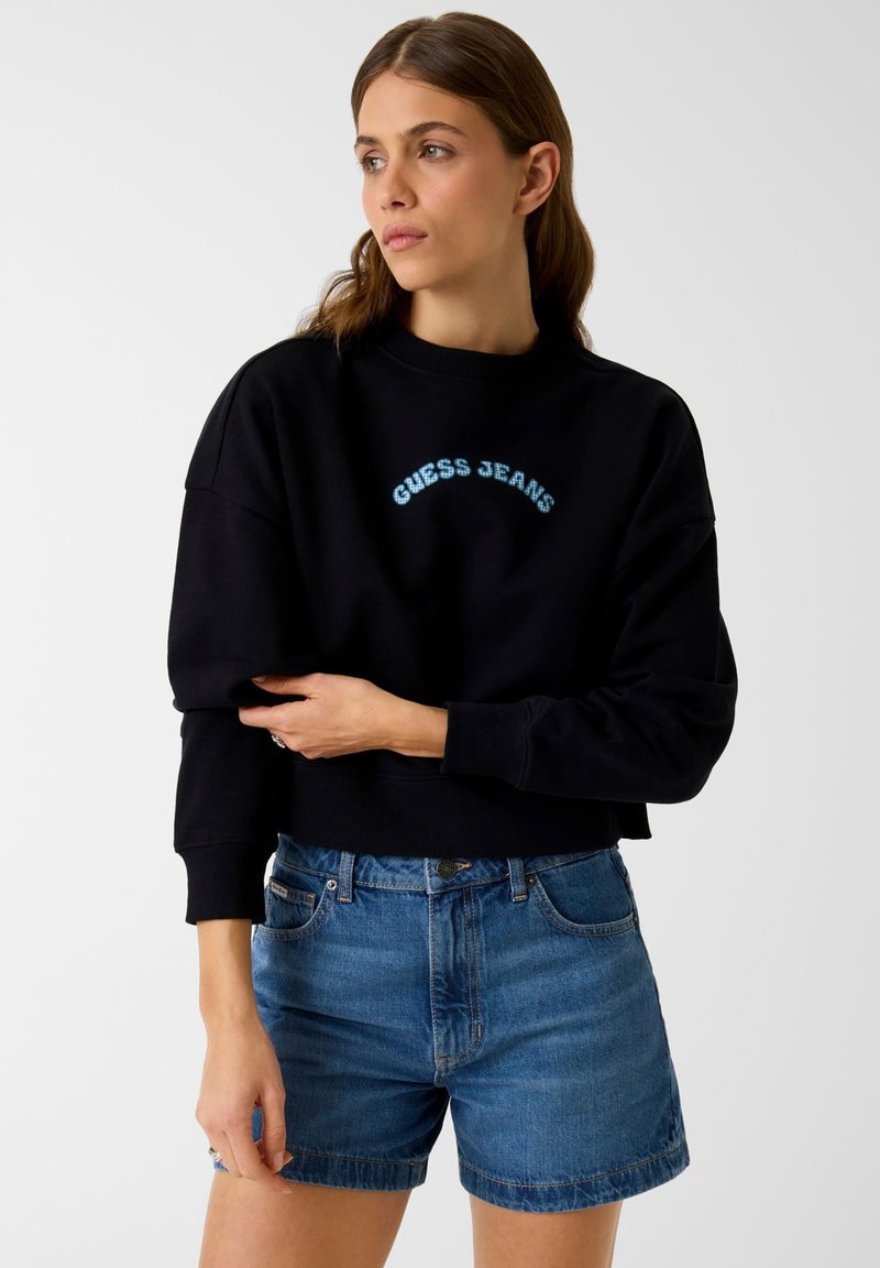 Nő fekete "GUESS JEANS" feliratú pulóverben és kék farmer rövidnadrágban, oldalra néz, kezeit a derekán összefonva egyszínű háttér előtt.