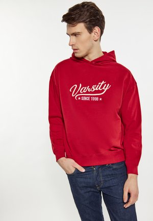 Młody mężczyzna w czerwonej bluzie z kapturem z napisem "Varsity Since 1998", niebieskie dżinsy, patrzy w lewo, jedną ręką w kieszeni.