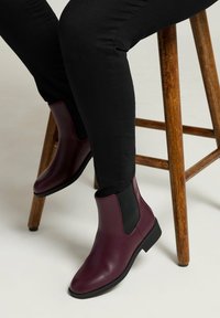 Bottines Chelsea en cuir bordeaux avec des panneaux latéraux élastiques, semelles plates et un design épuré, portées avec un jean noir, assis sur un tabouret en bois.