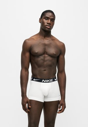 Atletisk mørkhudet mand, stående uden skjorte iført hvide Nike boxershorts mod en ensartet lys baggrund.