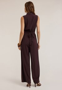 Salopetă fără mâneci, de culoare burgundă, cu guler și talie ajustabilă. Pantaloni larghi, de croială lejeră, dintr-un material neted și asortată cu brățări suprapuse.