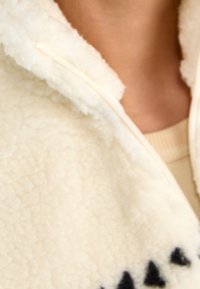 Smetanově bílá mikina z fleecu s vysokým límcem a zipem. Má texturovaný, nadýchaný povrch a černé trojúhelníkové vzory.