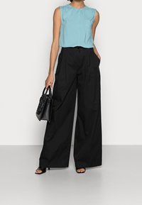 Blouse sans manches bleu clair avec une poche et pantalons larges noirs. Sac à main noir avec des accents texturés et talons ouverts noirs.