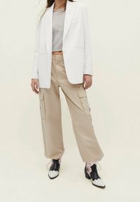 Femme portant un blazer blanc, un haut gris court, un pantalon cargo beige et des bottes de cowboy pointues noir et blanc, sur un fond uni.
