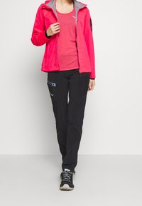 Giacca fucsia brillante con cerniera e dettagli grigi, abbinata a una canottiera rossa. Pantaloni neri con tasche e dettagli blu. Scarpe da ginnastica nere.