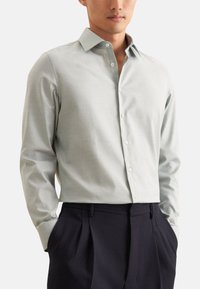 Camicia a maniche lunghe grigia chiara con colletto a bottoni, caratterizzata da un motivo testurizzato all'interno del colletto, abbinata a pantaloni scuri.