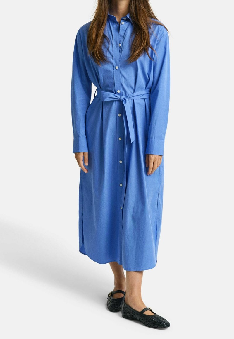 Femme portant une robe midi bleue à manches longues boutonnée, avec une ceinture nouée à la taille et des chaussures plates tressées noires, debout devant un fond uni.