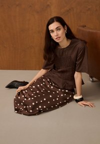 Femme aux longs cheveux foncés assise par terre portant un haut marron et une jupe marron à pois blancs, des bottes noires et deux bracelets.