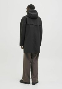 Jachetă impermeabilă neagră cu glugă, croială lejeră și detalii cu clapetă la spate. Asortată cu pantaloni maro largi și încălțăminte neagră.