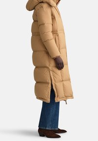 Lange, puffy jas in beige, met een capuchon en drukknoopsluitingen, gecombineerd met wijde blauwe jeans en bruine enkellaarsjes.