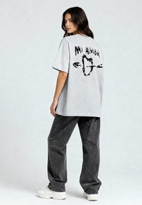 T-shirt oversize gris avec du texte graphique noir et des flèches dans le dos. Associé à un pantalon baggy gris foncé et des baskets blanches.
