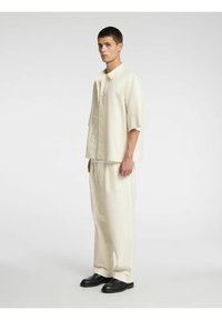 Beige kortermet skjorte med knappelukking foran, kombinert med ledige beige bukser og svarte slip-on sko. Minimalistisk design og nøytrale toner.