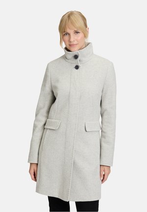 Frau in einem hellgrauen, knielangen Wollmantel mit hohem Kragen und zwei schwarzen Knöpfen, stehend vor einem schlichten, hellen Hintergrund.
