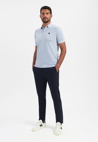 Lichtblauwe polo met een zwart logo op de borst, gecombineerd met donkerblauwe slim-fit broeken en witte sneakers. Soepele stof, casual ontwerp.