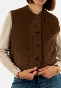 Gilet di velluto a coste marrone con scollatura rotonda, cinque bottoni e due grandi tasche frontali, indossato sopra un maglione leggero.