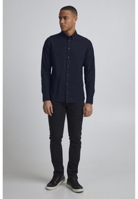 Marineblå skjorte med lange ermer, som har en subtil brodert logo, kombinert med svarte slim-fit bukser og svarte sneakers.