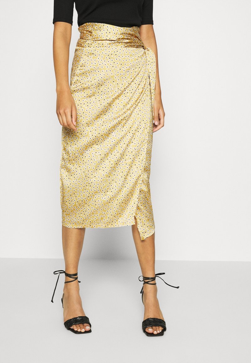 Femme portant une jupe midi jaune à fleurs avec une fente latérale, associée à des sandales à talons noires avec des brides et un haut noir.