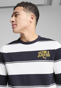 Prugasta majica s okruglim izrezom u mornarsko plavoj i bijeloj boji, sa teksturiranim zlatnim logotipom "PUMA sports club" na prsima. Mekana, ležerna tkanina.