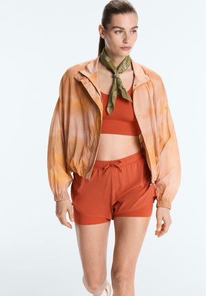 Kvinde iført en gennemsigtig orange tie-dye jakke, rustfarvet sports-bh, matchende shorts og et grønt mønstret halstørklæde, stående mod en hvid baggrund.