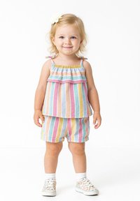 Ett ungt barn står och ler, klädd i en pastellfärgad randig topp och shorts, vita strumpor, blommiga sneakers och en vit hårklämma.