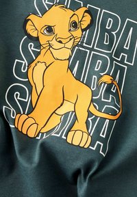 T-shirt en tissu vert avec un lion de dessin animé orange aux grands yeux et contours noirs. Texte "SIMBA" en blanc affiché derrière le lion.