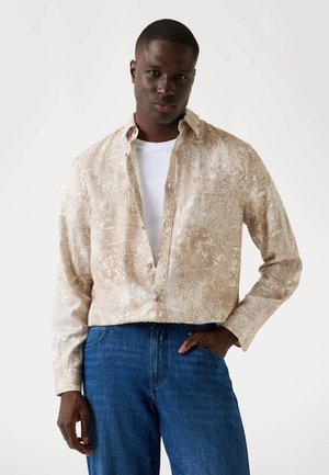 Short-sleeve shirt in beige fabric with white floral patterns, relaxed fit, and button-down collar, paired with blue denim jeans.