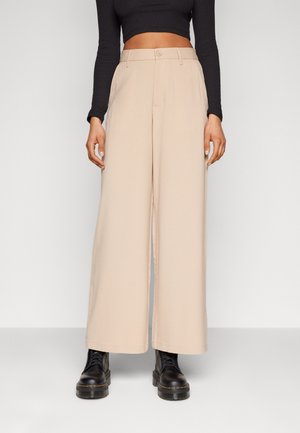 Béžové široké kalhoty nošené s černým krátkým topem s dlouhými rukávy a černými masivními šněrovacími botami na neutrálním pozadí.