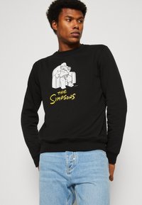 Sudadera negra con un gráfico de dibujo animado de un personaje sentado en una silla, texto "Los Simpson" en amarillo, puños y dobladillo acanalados.