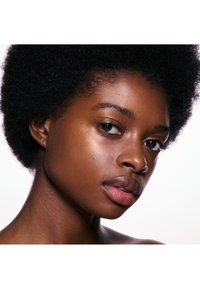 Gros plan d'une jeune femme noire avec des cheveux afro naturels, une peau lisse et une pierce au nez, regardant légèrement sur le côté devant un fond blanc.