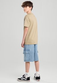 T-shirt corto beige con maniche corte, vestibilità ampia, abbinato a pantaloni cargo azzurri chiari con tasche laterali. Sneakers nere con dettagli bianchi.