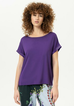 Frau mit lockigem Haar, die ein lockeres lila Kurzarmoberteil und mehrfarbige Blumenhose vor einem schlichten hellen Hintergrund trägt.