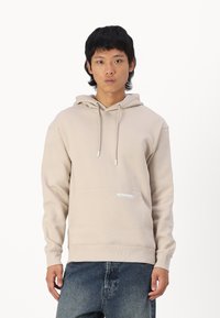 Hoodie beige in cotone misto, dotato di tasca frontale, cappuccio con cordino e etichetta bianca in basso a sinistra.