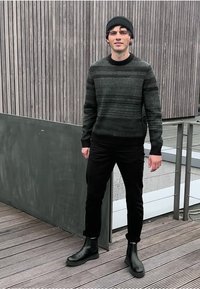 Svarta ankelstövlar, mörka jeans och en mörkgrön randig tröja. Designen har en rund halsringning och ribbade mudd. Står på träterrass.