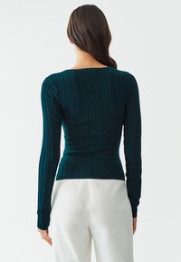 Dark teal long-sleeve top with a textured pattern, fitted design, and rounded neckline. Smooth fabric with ribbed accents along the sleeves.