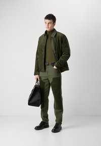 Green corduroy jacket over olive turtleneck, matching green trousers with textured patches, black leather shoes, and a black handbag.