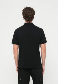 Άνδρας με σγουρά μαλλιά που φ�οράει ένα απλό μαύρο κοντομάνικο πόλο και μαύρο παντελόνι cargo, εικονιζόμενος από πίσω σε λευκό φόντο.