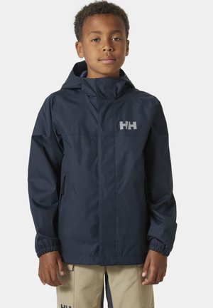 Niño con chaqueta azul marino con capucha y logo en el pecho, y pantalones beige, de pie contra un fondo claro y uniforme.