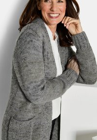 Lächelnde Frau mit braunen Haaren, die eine graue Strickjacke über einem weißen Shirt trägt und sich an einer weißen Fläche vor einem schlichten Hintergrund lehnt.