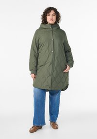 Olivzaļš ķeburains mētelis ar kapuci, priekšpusē ar spiedpogām un sānu kabatām, valkāts kopā ar zilām plaša kāju biksēm un brūniem apaviem.